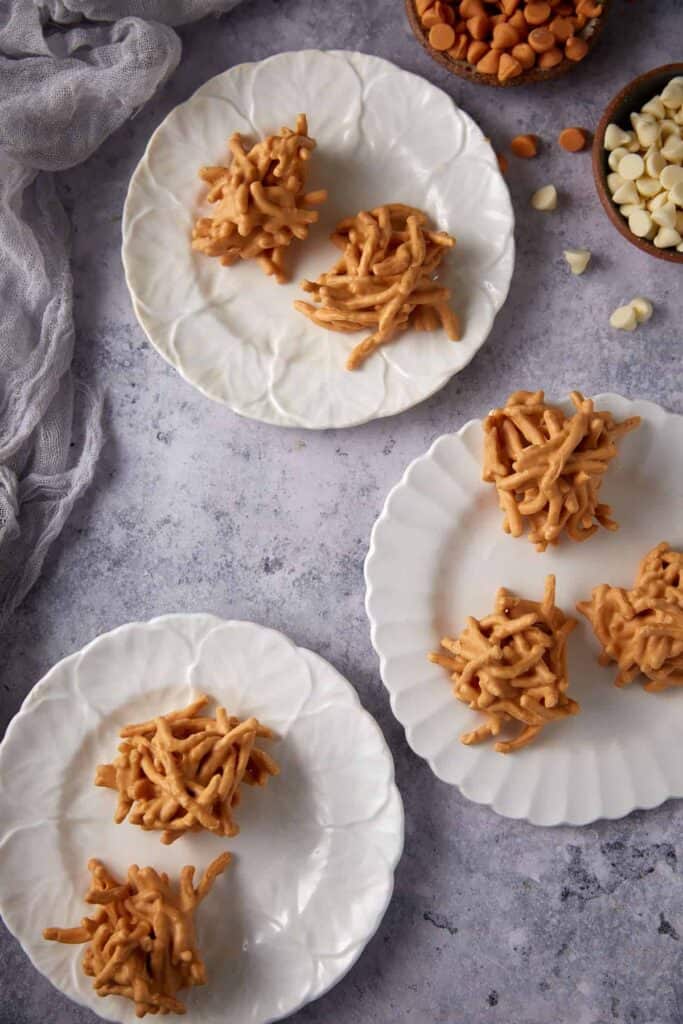 Four ingredient haystack cookies served on white plates with bowls of white chocolate and butterscotch chips nearby.