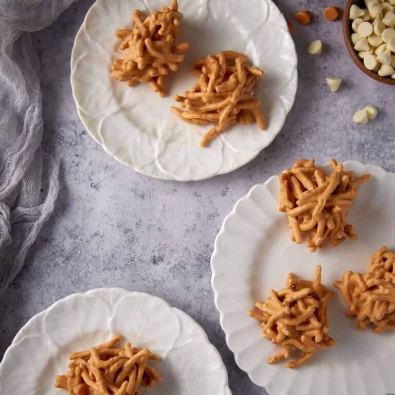 Four ingredient haystack cookies served on white plates with bowls of white chocolate and butterscotch chips nearby.