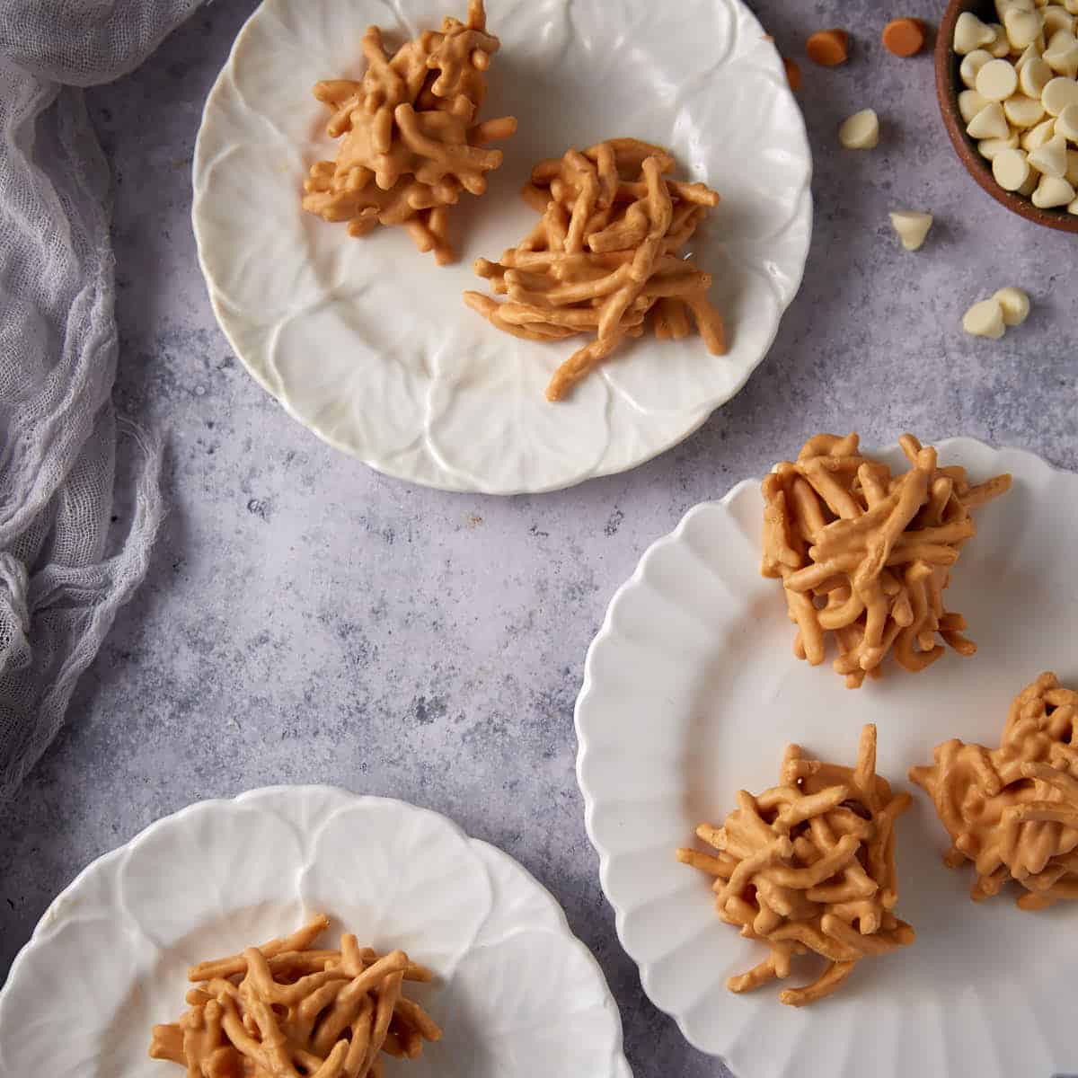 Four ingredient haystack cookies served on white plates with bowls of white chocolate and butterscotch chips nearby.