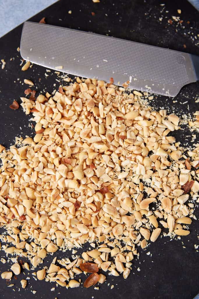 Chopped roasted peanuts on a cutting board with a chef’s knife.