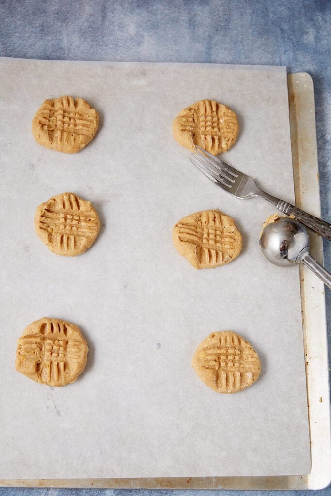 Peanut butter cookie dough scooped onto a parchment-lined baking sheet and pressed with a fork into a crisscross pattern.