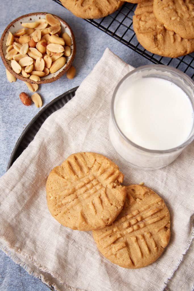 Four peppery peanut butter cookies on a linen napkin with a glass of milk, peanuts, and a cooling rack of cookies behind them.