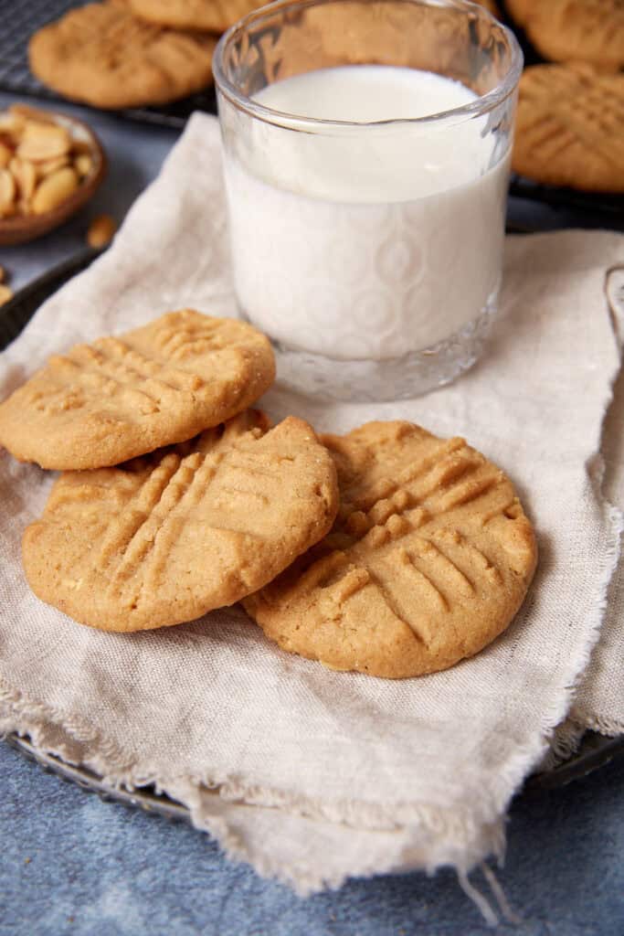 Three peppery peanut butter cookies on a linen napkin with a glass of milk, peanuts, and a cooling rack of cookies behind them.