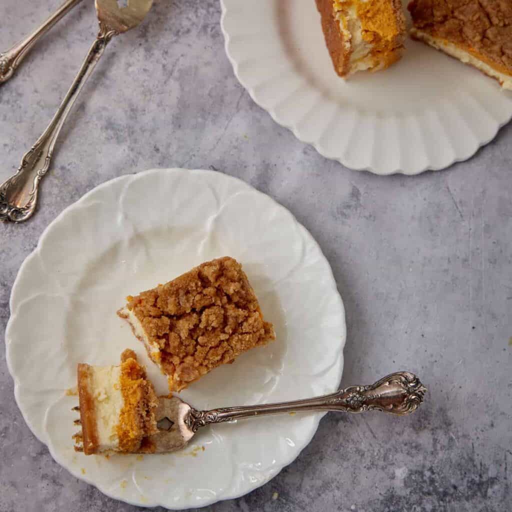 Pumpkin pie cheesecake bars served on white plates with a forkful showing the creamy double cheesecake layers and crumb topping.