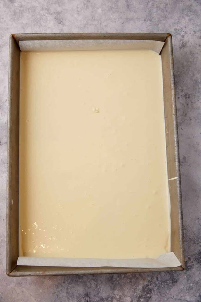 Plain cheesecake filling spread in a parchment-lined 9x13 pan for the first layer of pumpkin pie cheesecake bars.