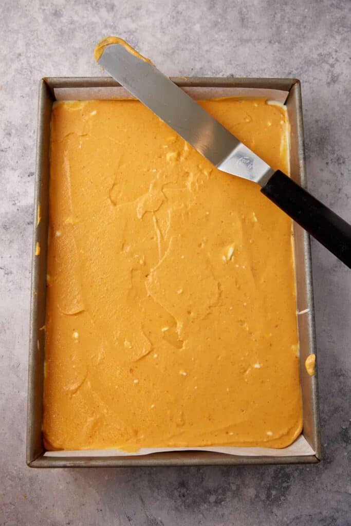 Pumpkin cheesecake filling spread over the first cheesecake layer in a parchment-lined baking pan.