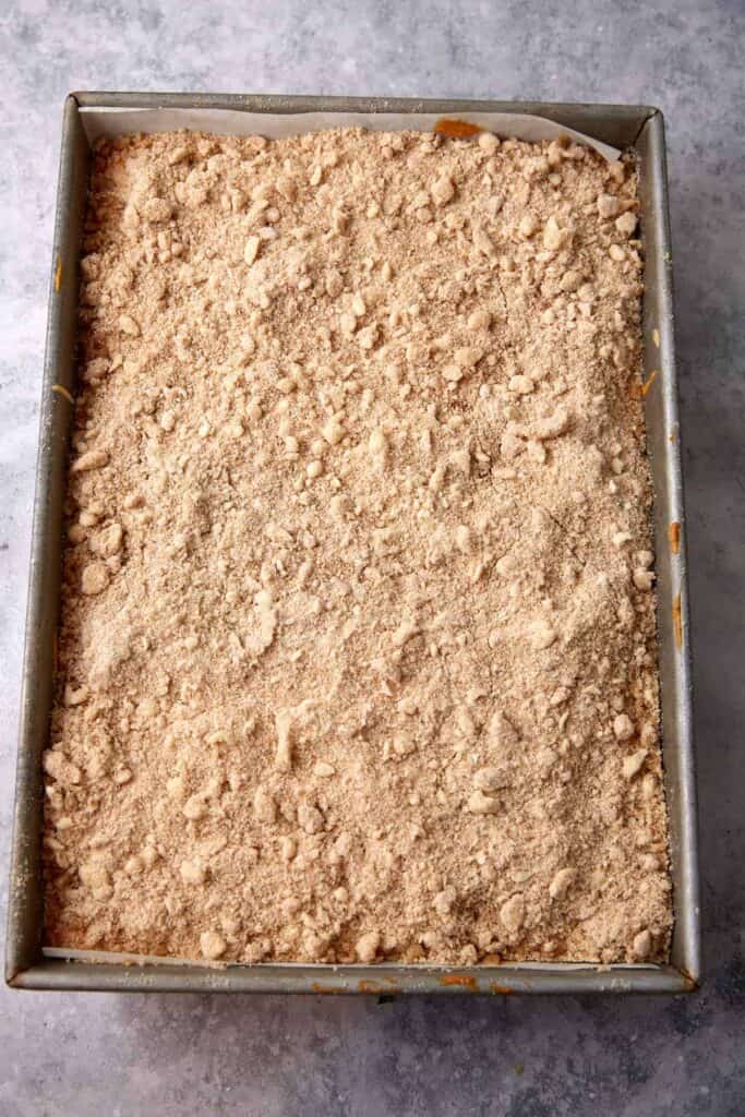 Unbaked pumpkin pie cheesecake bars in a parchment-lined pan covered with an even layer of spiced crumble topping.