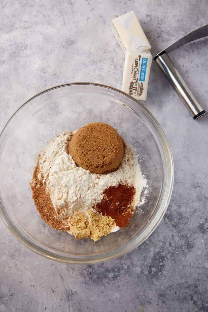 Flour, brown sugar, cinnamon, ginger, and nutmeg in a glass bowl with butter nearby for the crumble topping.