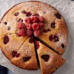 Square crop of raspberry ricotta cake dusted with powdered sugar and topped with fresh raspberries, with one slice removed.