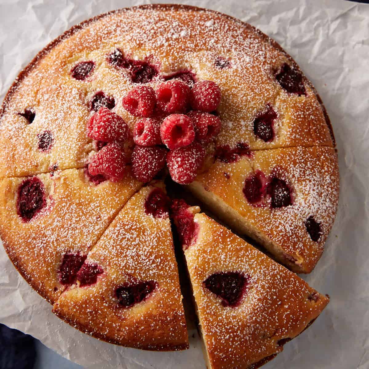 Square crop of raspberry ricotta cake dusted with powdered sugar and topped with fresh raspberries, with one slice removed.