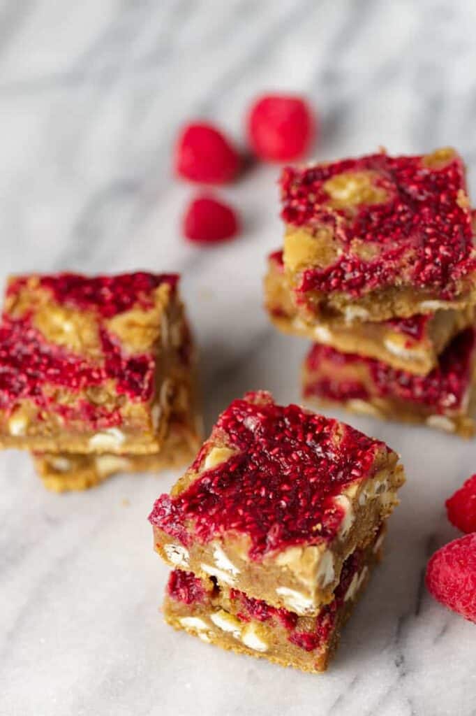 Raspberry white chocolate blondie bars stacked on a marble surface with fresh raspberries alongside.