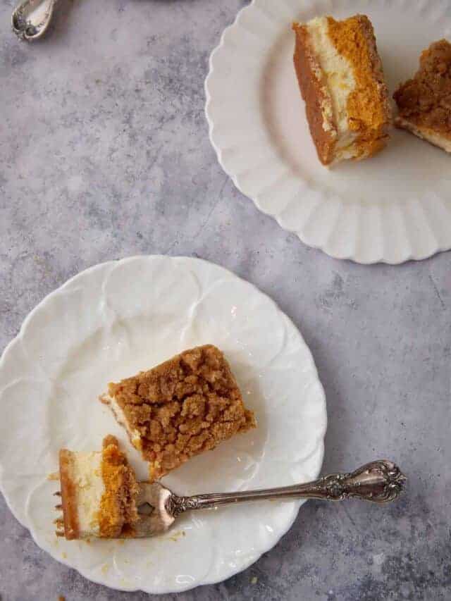 Pumpkin pie cheesecake bars served on white plates with a forkful showing the creamy double cheesecake layers and crumb topping.