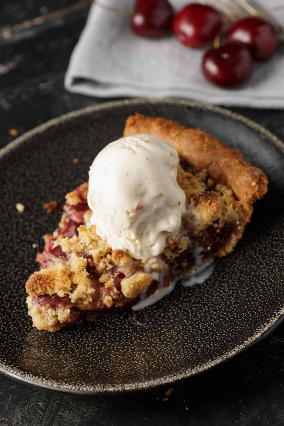 Fresh Cherry Pie with Crumb Topping served on a black plate with white specks, with a scoop of vanilla ice cream on top.