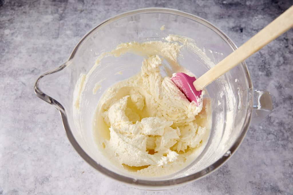 Butter and sugar creamed together in a glass stand mixer bowl, pale and fluffy, with a pink silicone spatula resting in the bowl.