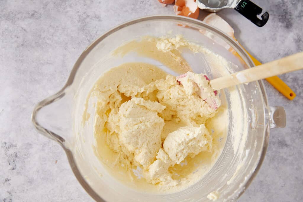 Yellow cake batter in a glass stand mixer bowl after eggs have been incorporated, with a pink spatula and empty egg shells visible in the background.