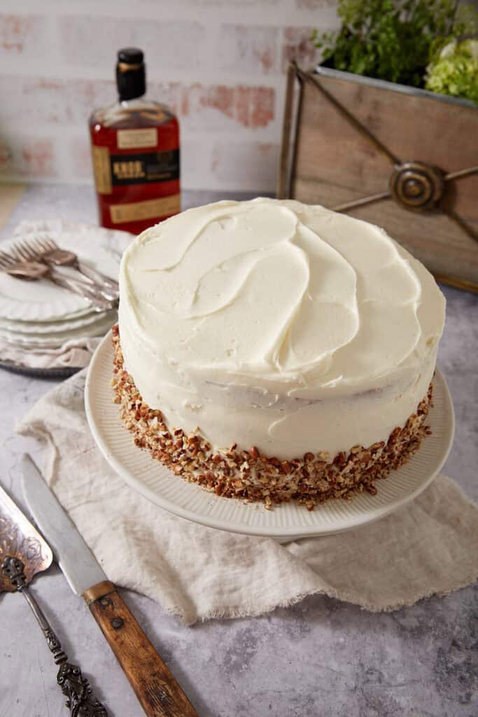 Whole Pecan Pie Cake frosted with bourbon cream cheese frosting on a white cake stand, chopped pecans pressed around the bottom edge, with a bottle of Knob Creek bourbon and stacked white plates in the background.