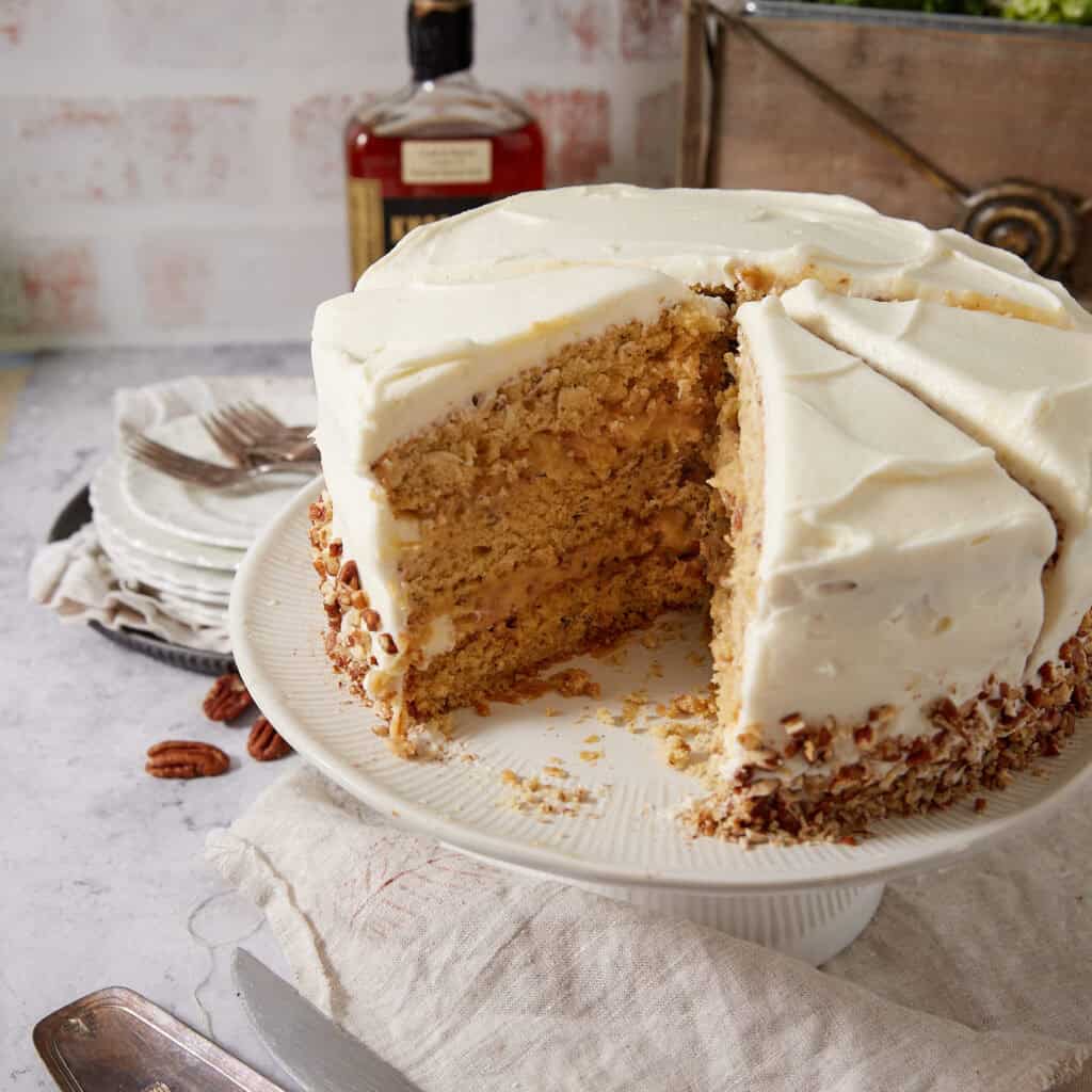 Pecan Pie Cake on a white cake stand with two slices cut and one removed, revealing the layers of pecan cake and the pale gold pecan pie filling, with a bourbon bottle and stacked plates in the background.