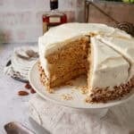 Pecan Pie Cake on a white cake stand with two slices cut and one removed, revealing the layers of pecan cake and the pale gold pecan pie filling, with a bourbon bottle and stacked plates in the background.