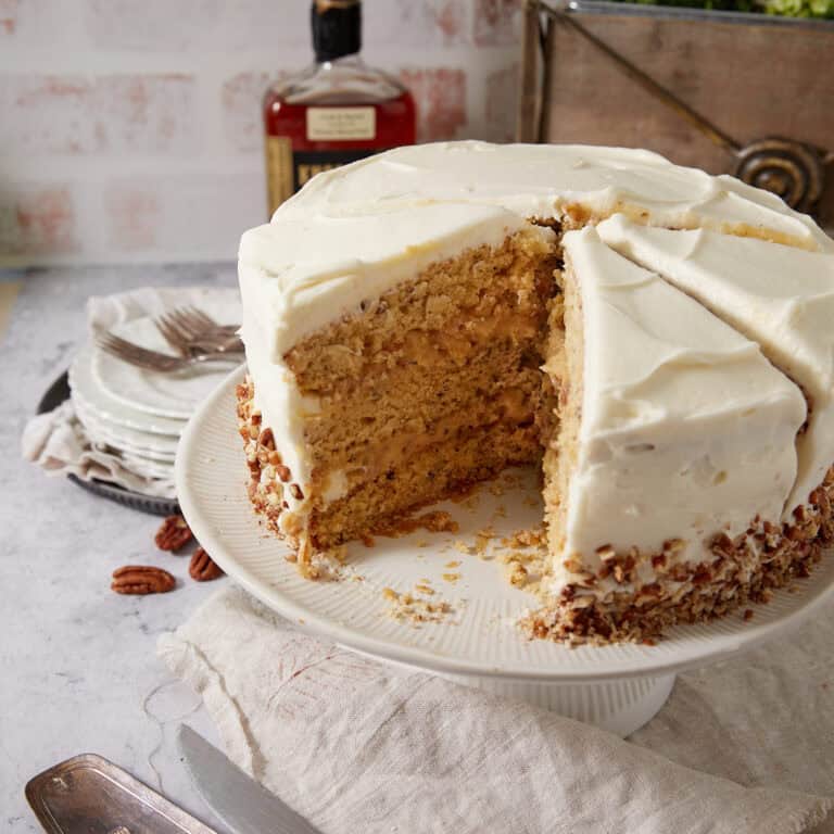Pecan Pie Cake on a white cake stand with two slices cut and one removed, revealing the layers of pecan cake and the pale gold pecan pie filling, with a bourbon bottle and stacked plates in the background.