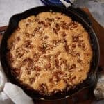 Overhead square crop of the finished pecan pie cobbler in a cast iron skillet on a wooden trivet, golden batter risen and cracked on top with chopped pecans throughout, with a white linen and silver spoon alongside.
