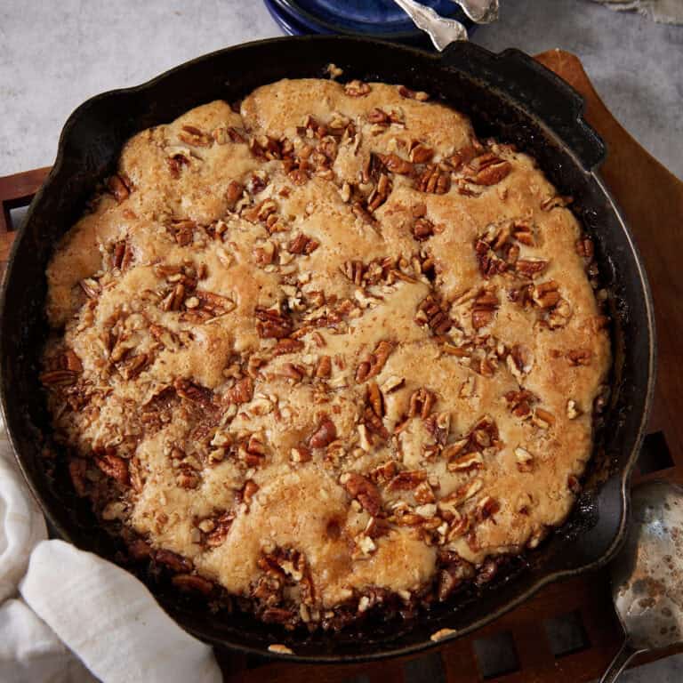 Overhead square crop of the finished pecan pie cobbler in a cast iron skillet on a wooden trivet, golden batter risen and cracked on top with chopped pecans throughout, with a white linen and silver spoon alongside.