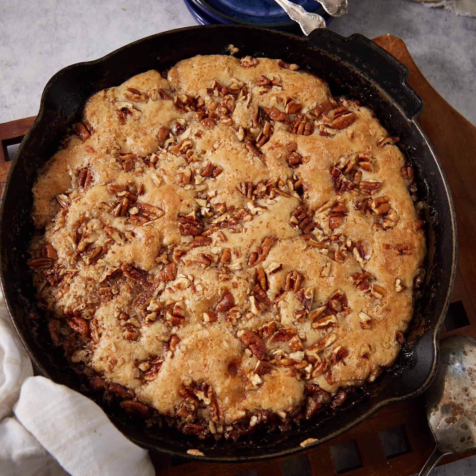 Overhead square crop of the finished pecan pie cobbler in a cast iron skillet on a wooden trivet, golden batter risen and cracked on top with chopped pecans throughout, with a white linen and silver spoon alongside.