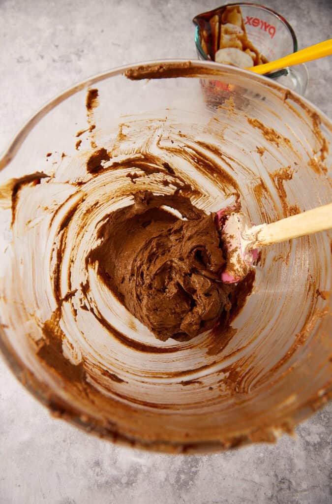 Cookie dough in a glass mixing bowl after the molasses has been beaten in, dark and glossy, with a pink spatula and a measuring cup of molasses in the background.