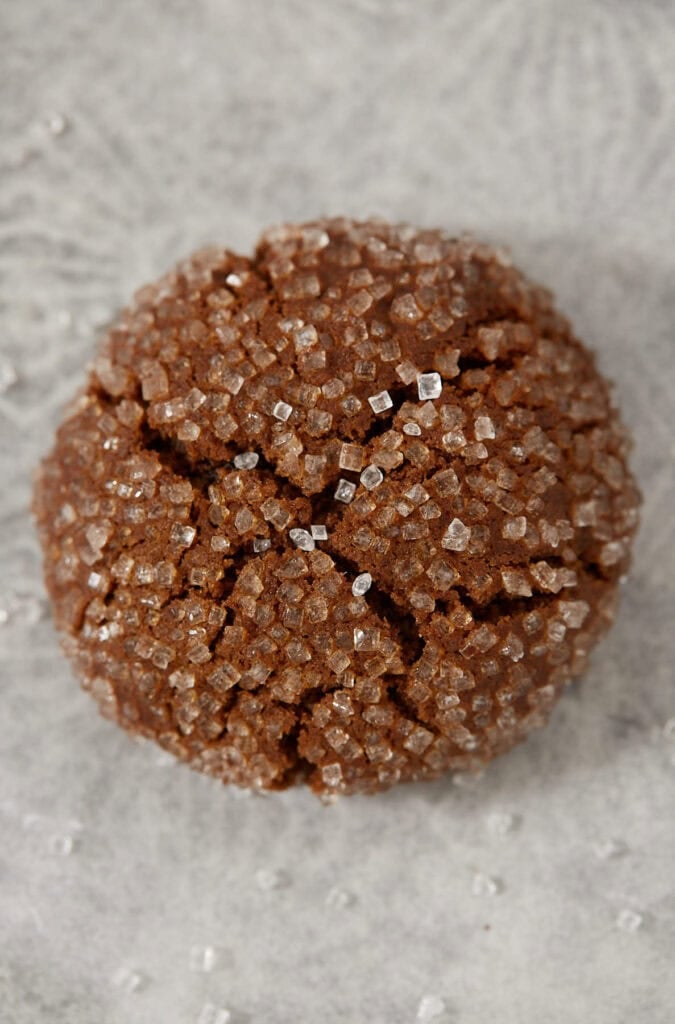 Overhead view of a single molasses crinkle cookie coated in coarse sparkling sugar with a crackled top, on a gray patterned surface.