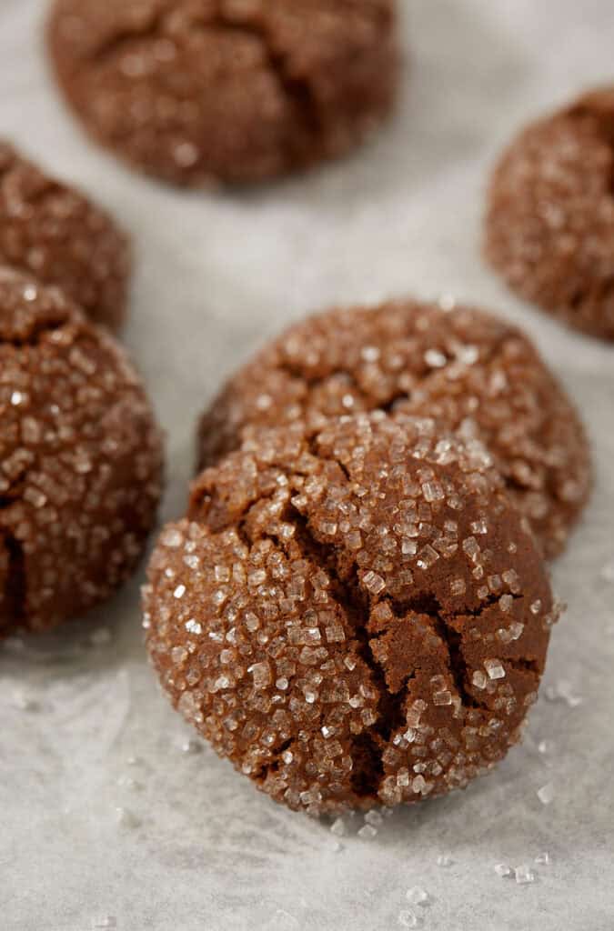 A molasses crinkle cookie standing on parchment paper with more cookies behind it, showing the crackled sugar crust and loose sugar crystals scattered around.