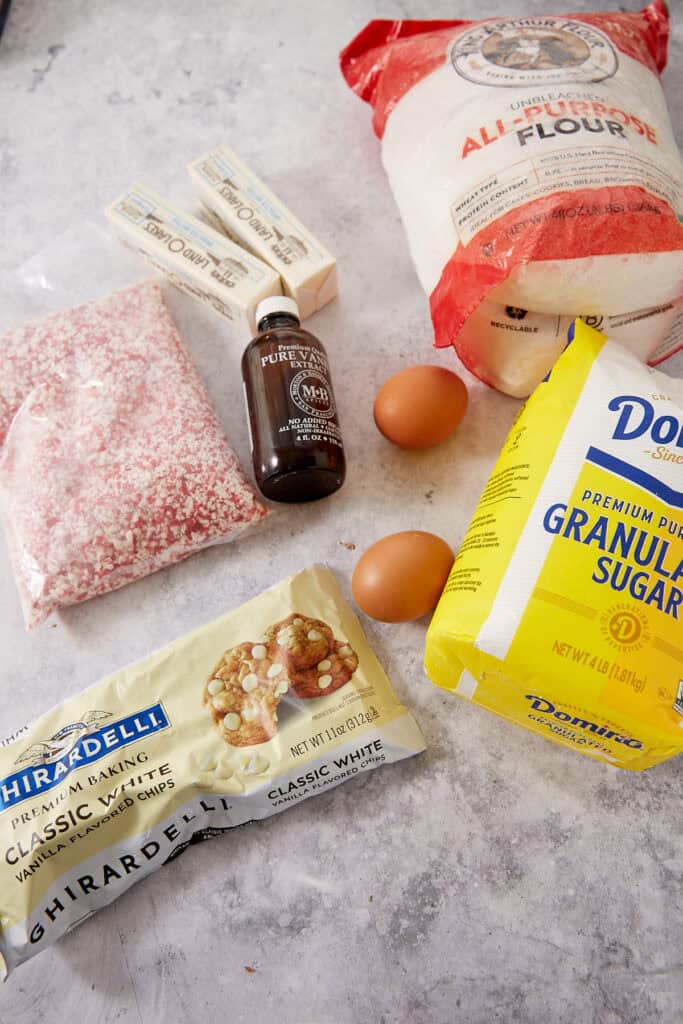 Ingredients for baking arranged on a counter: a bag of flour, a bag of sugar, white chocolate chips, peppermint bits, two sticks of butter, two eggs, and vanilla extract.