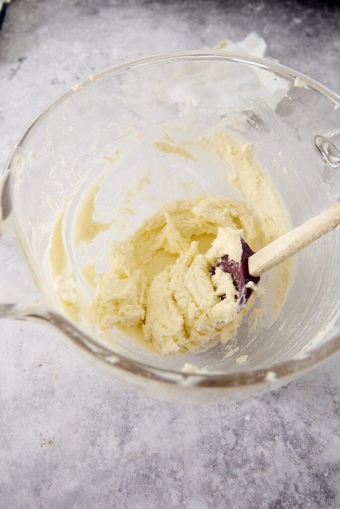 A purple spatula mixing pale cookie dough in a clear glass bowl.