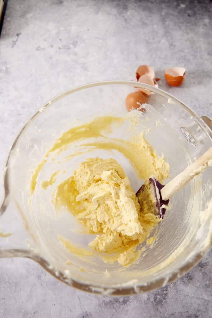 A glass mixing bowl containing creamed butter and sugar, with a spatula and eggshells nearby.