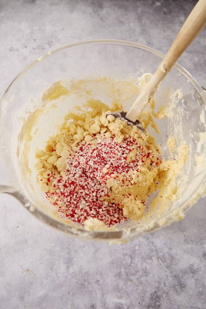 A wooden spatula stirs white chocolate chips and crushed peppermint candies into cookie dough inside a glass mixing bowl.