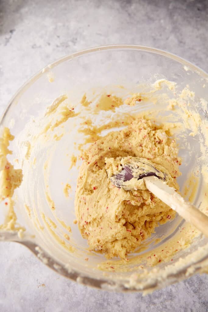Cookie dough with red flecks in a glass mixing bowl with a rubber spatula.