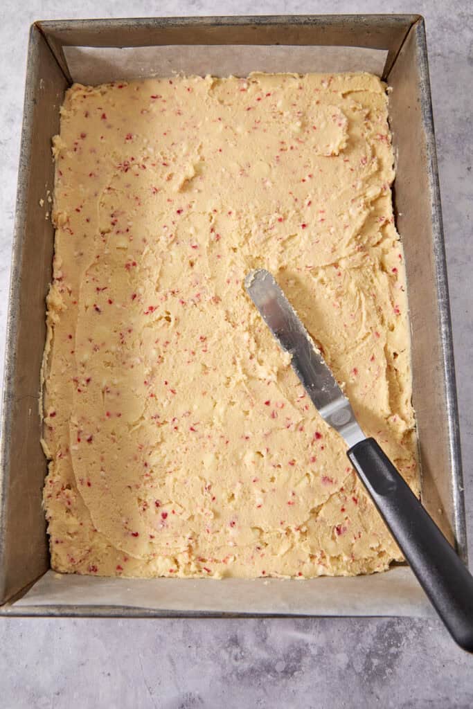 A metal spatula spreading cookie dough with red specks into a parchment-lined rectangular baking pan.