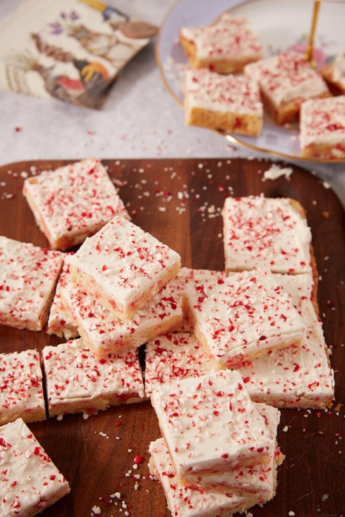 Peppermint bark cookie bars with white frosting and crushed candy cane topping, arranged on a wooden cutting board.