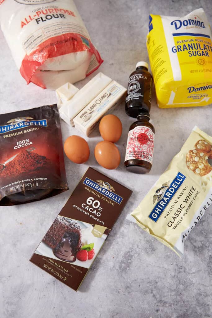 Baking ingredients on a counter, including a bag of flour, a bag of sugar, two sticks of butter, three eggs, cocoa powder, a bar of bittersweet chocolate, white chocolate chips, vanilla extract, and peppermint extract.