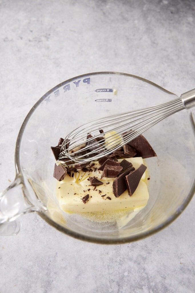 A glass measuring cup containing sticks of butter and chopped dark chocolate pieces with a metal whisk resting on top.