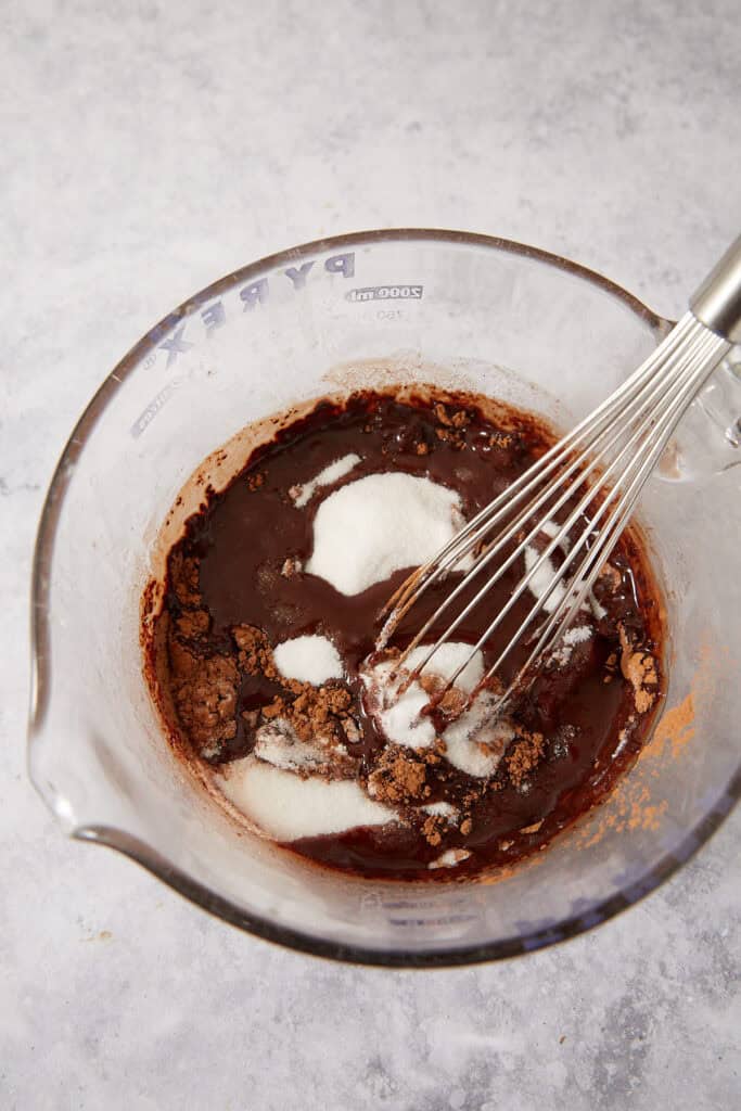 A wire whisk mixing white sugar and cocoa powder into a dark liquid in a glass measuring cup.