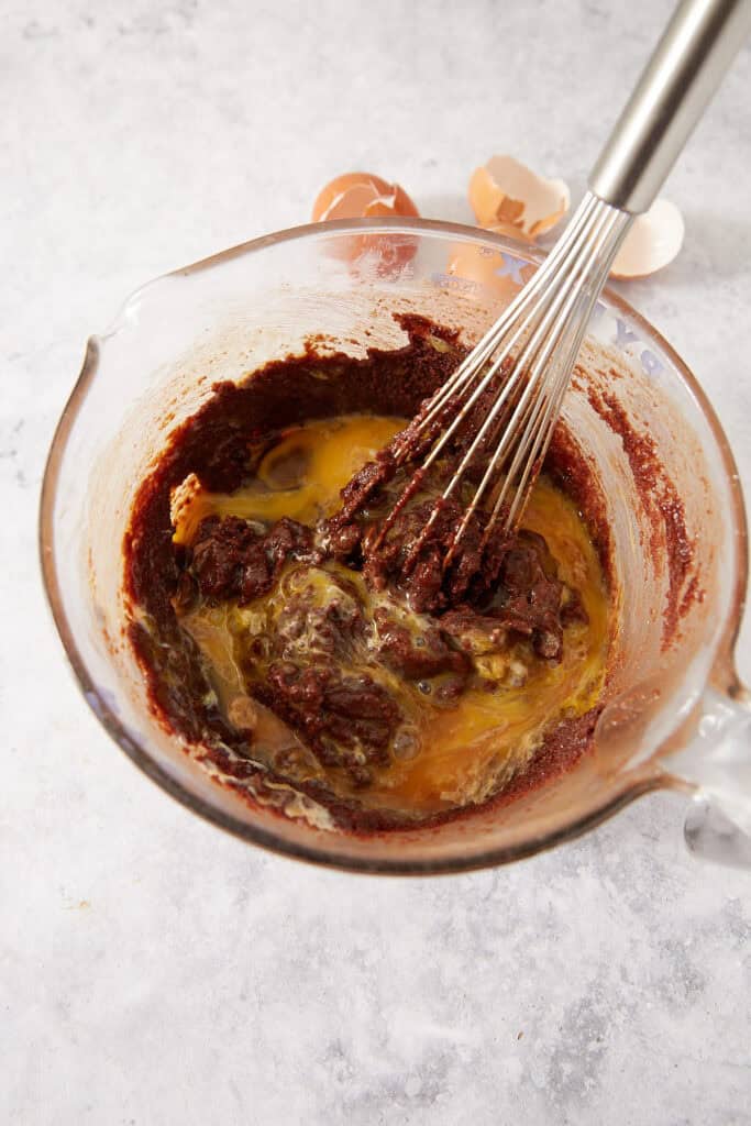 A wire whisk blending raw eggs into dark chocolate batter in a glass measuring cup.