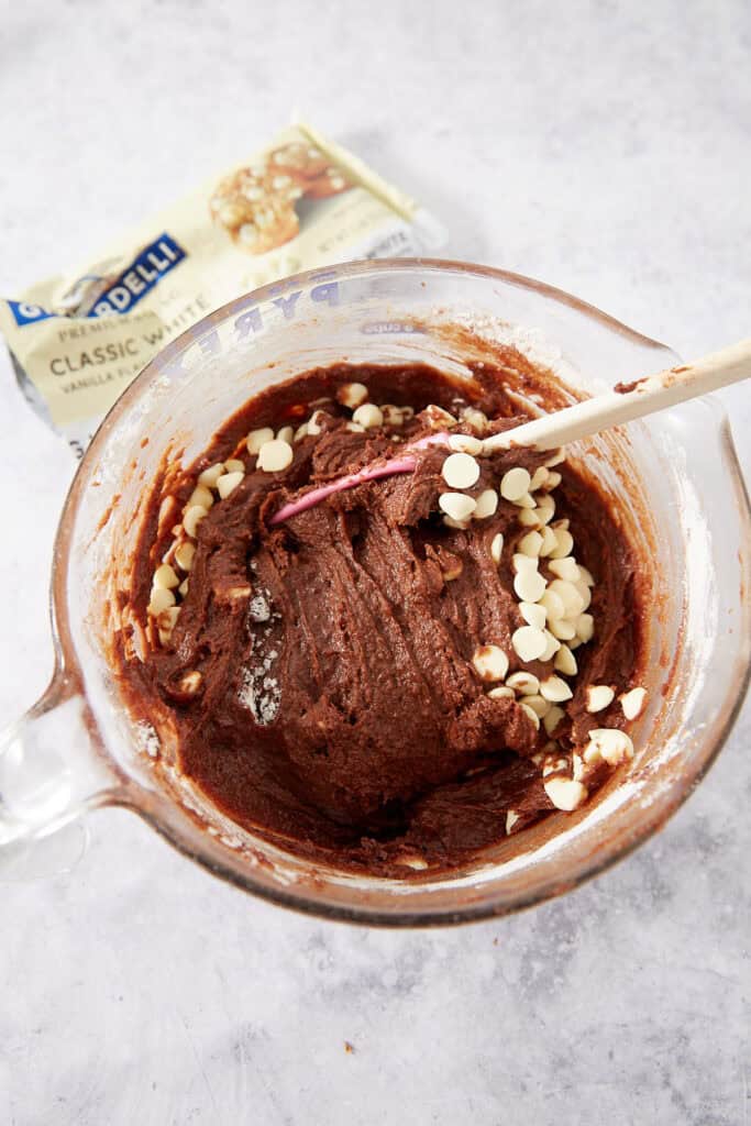 A spatula folds white chocolate chips into dark chocolate cookie dough in a glass mixing bowl, with a bag of white chocolate chips in the background.