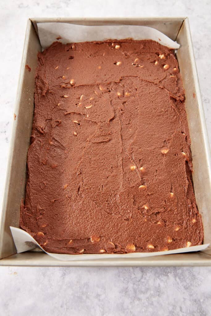 Unbaked chocolate brownie batter with white chips spread in a rectangular baking pan lined with parchment paper.