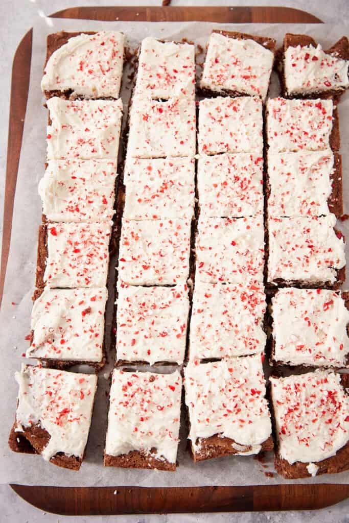 A grid of square chocolate brownies topped with white frosting and crushed peppermint candies on a wooden board.
