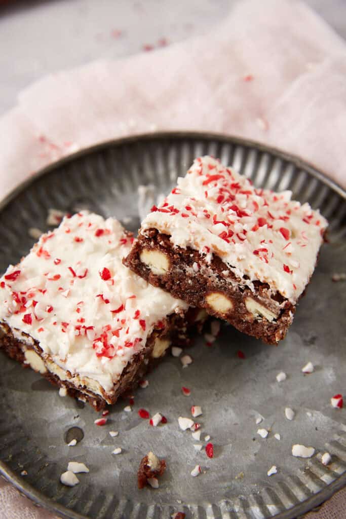 Two square chocolate brownies topped with white frosting and crushed peppermint candy on a metal plate.