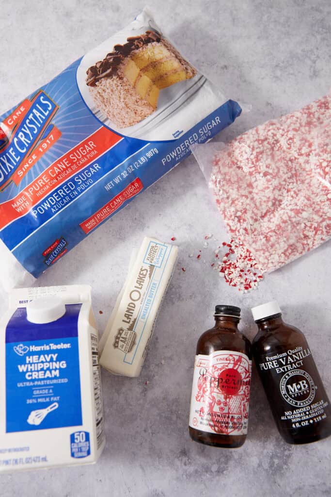 Ingredients for a dessert recipe: a bag of Dixie Crystals powdered sugar, a pile of crushed peppermint, a stick of Land O Lakes unsalted butter, a carton of heavy whipping cream, and bottles of peppermint and vanilla extract.