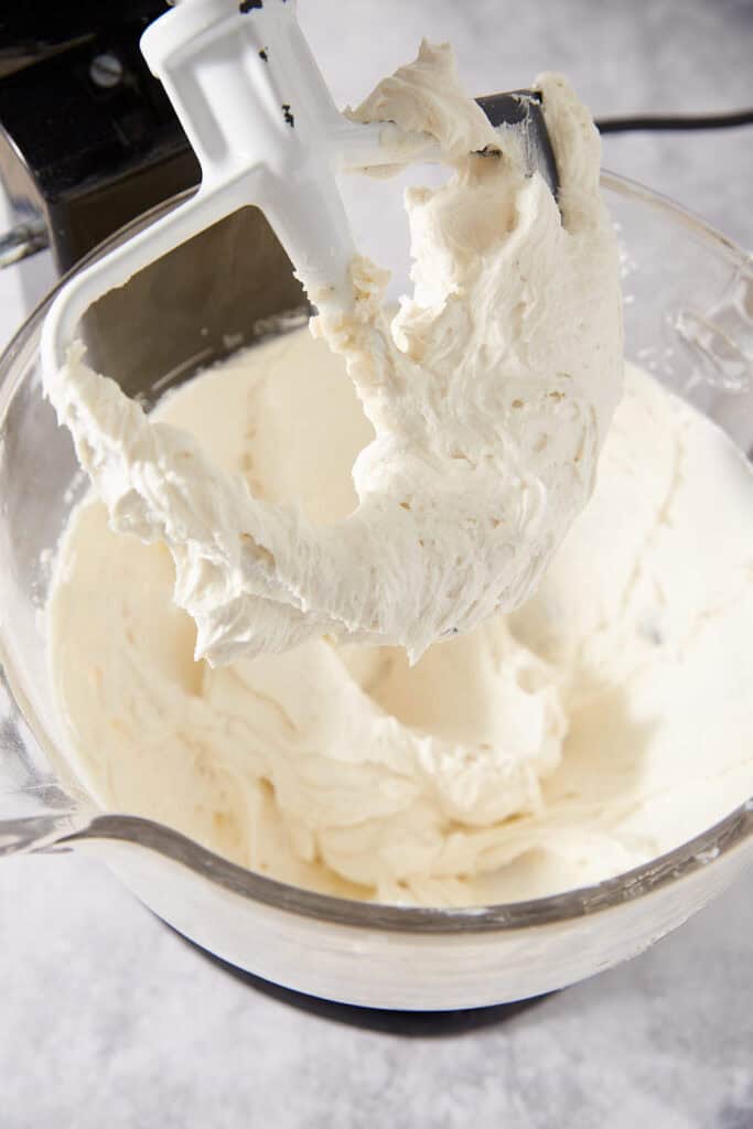 A flat beater attachment covered in thick, pale cream frosting inside a glass mixing bowl.