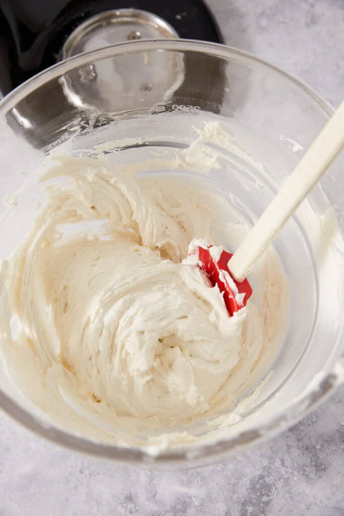 A red silicone spatula mixing white frosting in a glass bowl.